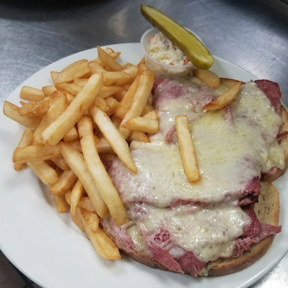 Open-faced sandwich topped with melted cheese and sliced meat, served with French fries, coleslaw, and a pickle on a white plate.