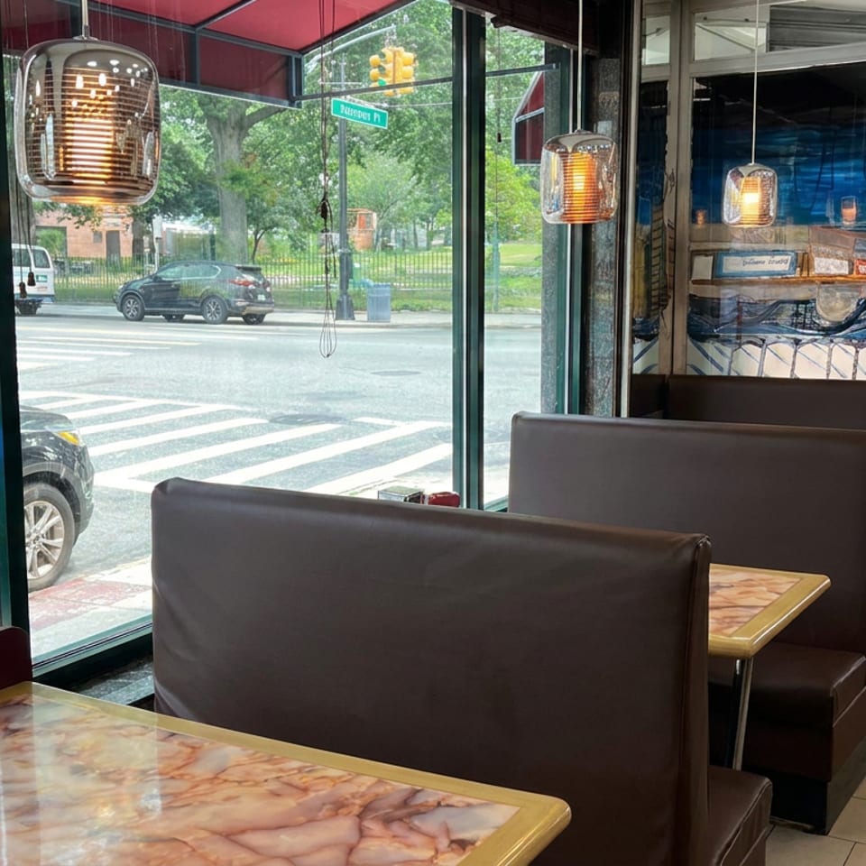 Diner interior with leather booths, marble tables, pendant lights, and large windows overlooking a city intersection and park.