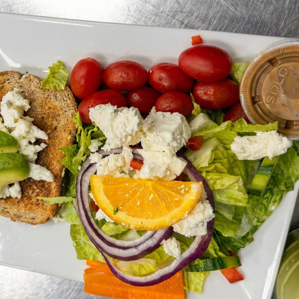 Greek-style salad with lettuce, tomatoes, feta, avocado, orange, onions, toasted bread, and dressing on a white plate.